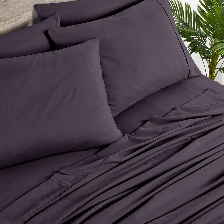 Purple bedding set with pillows on a bed, featuring a plant in the background.