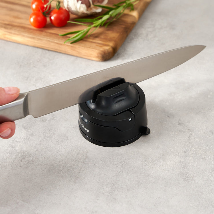 Knife being sharpened on a black sharpener with a cutting board and vegetables in the background.