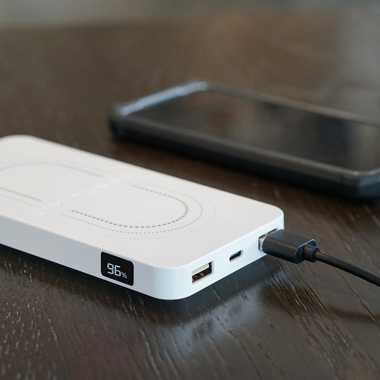 White power bank with digital display on a wooden surface, connected to a smartphone.