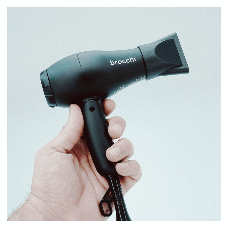 Hand holding a black hair dryer with 'brocchi' branding against a light blue background