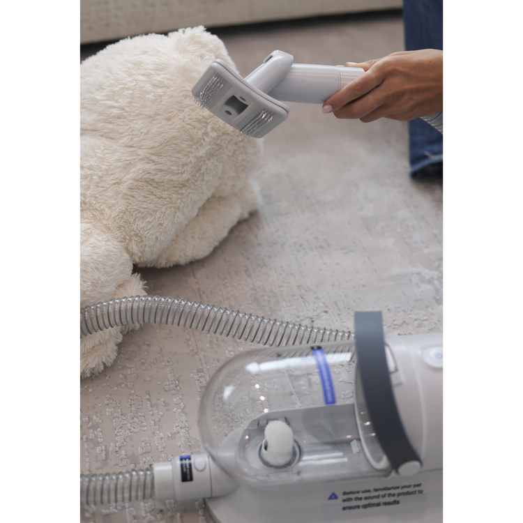 Person using a handheld device on a fluffy white dog with a vacuum cleaner on the floor.