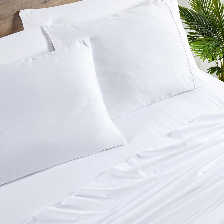 White bed sheets and pillows on a bed with a plant in the corner.