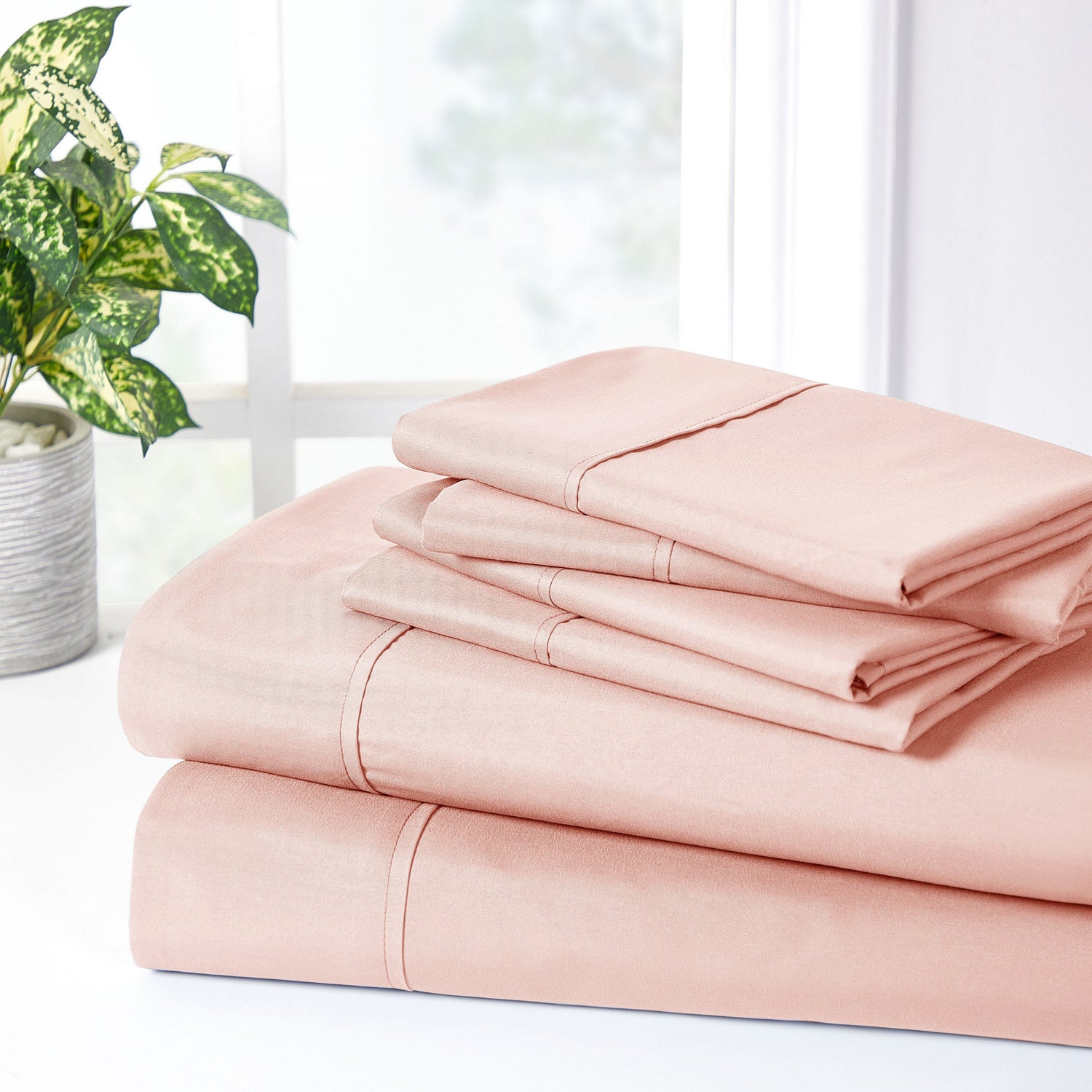 Stack of pink bed sheets on a white surface with a plant in the background