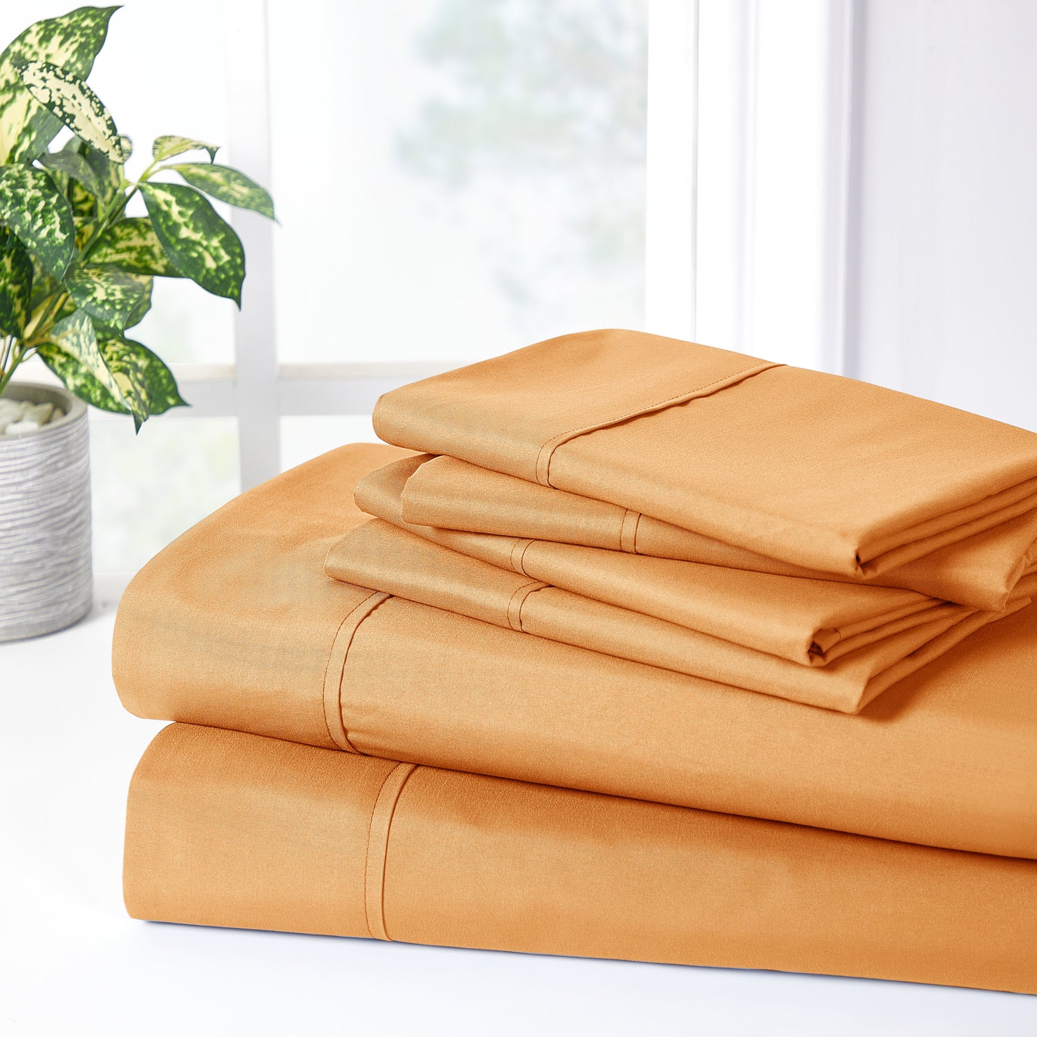Orange bed sheets stacked on a white surface with a plant in the background