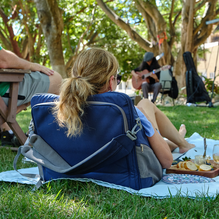 Person sitting on a blanket with a blue backpack, surrounded by trees and people in a park setting.