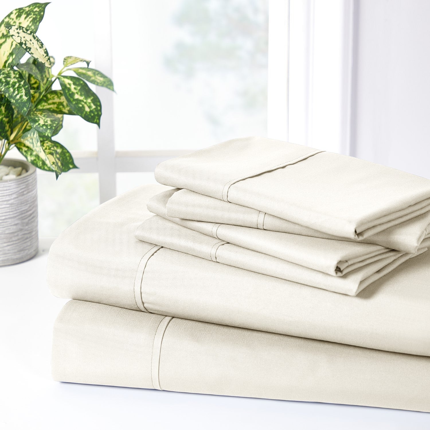 Stack of beige bed sheets on a white surface with a plant in the background