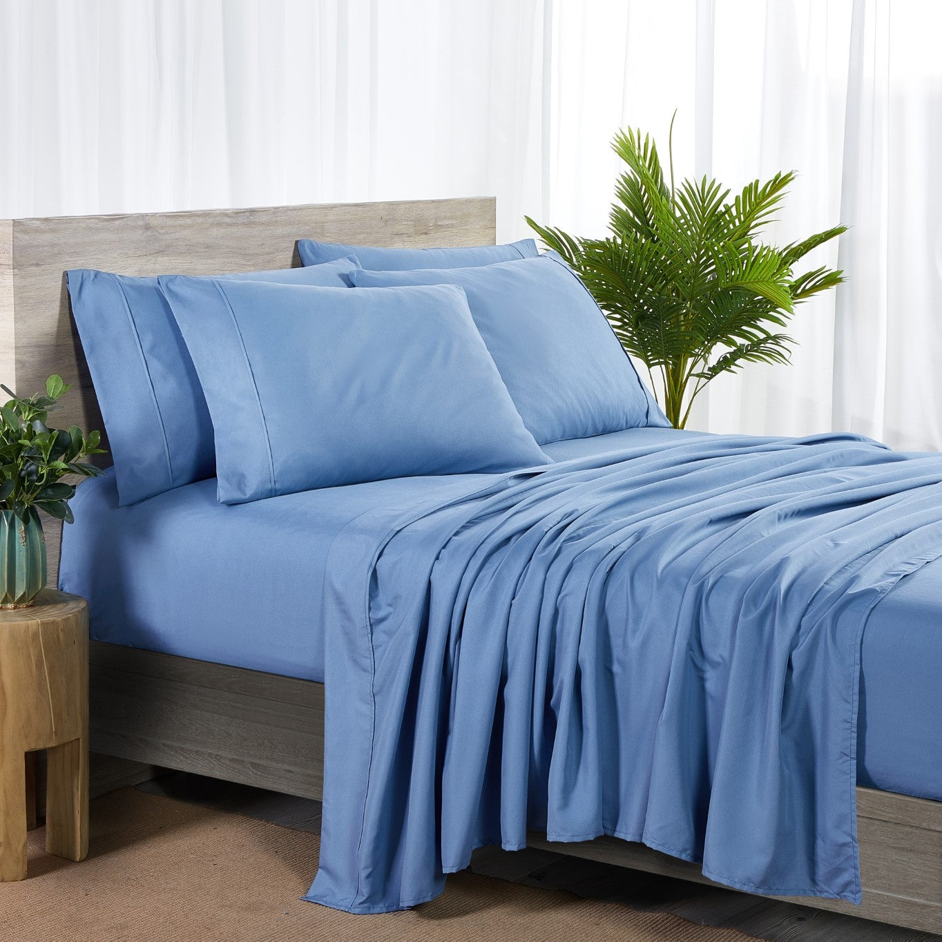 Blue bedding set on a bed with a wooden headboard and plants in the background.