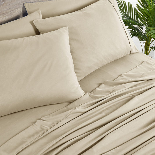 A warm taupe colored 6-piece bamboo sheet set on a bed, with a flat sheet, fitted sheet, and pillowcases displayed. The bed is set against a room backdrop with a plant and nightstand on the side.