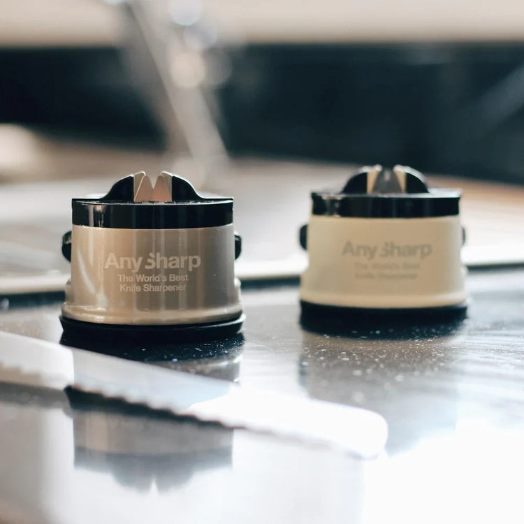 Two 'Any Sharp' knife sharpeners on a reflective surface with a blurred background.