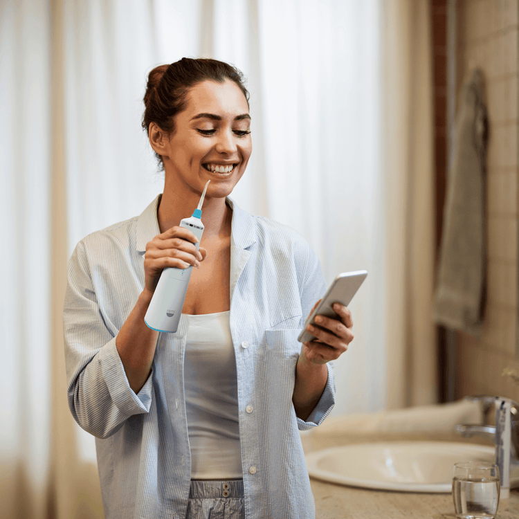 Woman using a dental water flosser and holding a smartphone in a bathroom.
