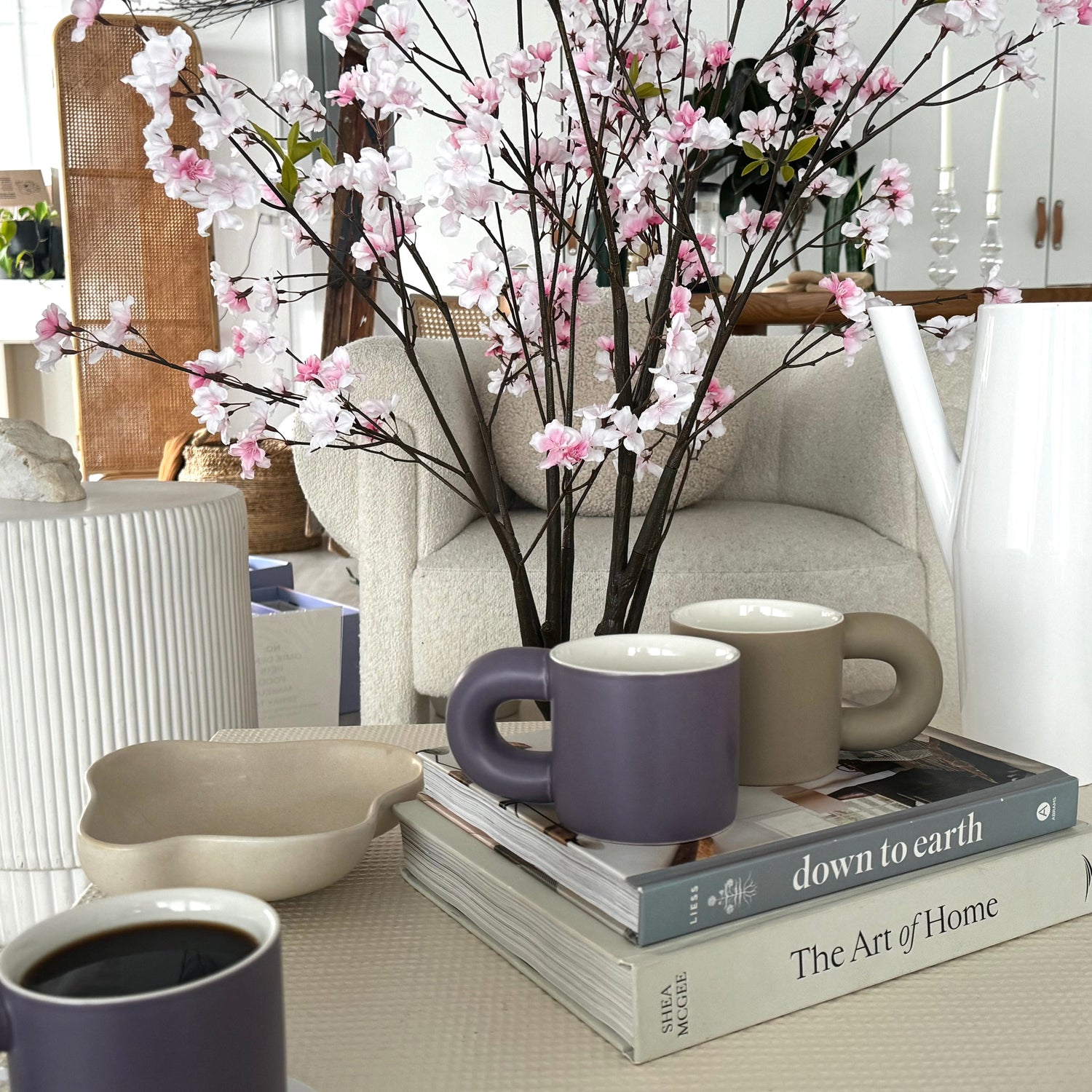The image features a cozy setting with two ceramic mugs, one purple and one gray, placed on a table alongside a heated coaster and a silver spoon. The scene includes a decorative vase with pink cherry blossoms, adding a touch of elegance and tranquility to the atmosphere.