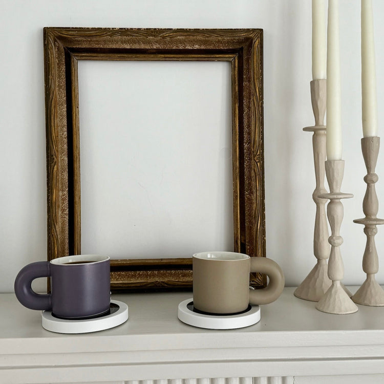 Two mugs on coasters with a framed picture and candlesticks in the background.