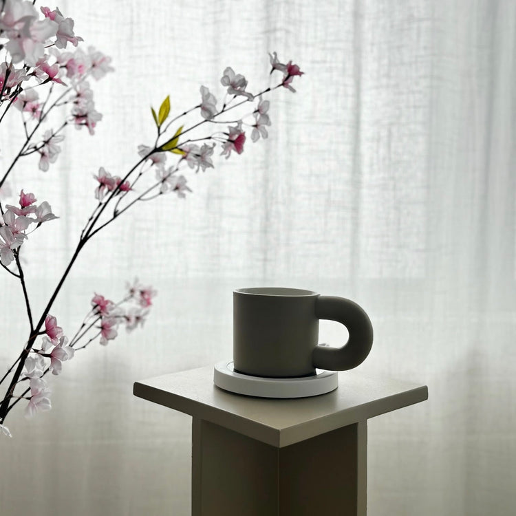 Gray mug on a white coaster with cherry blossom branches against a white curtain background