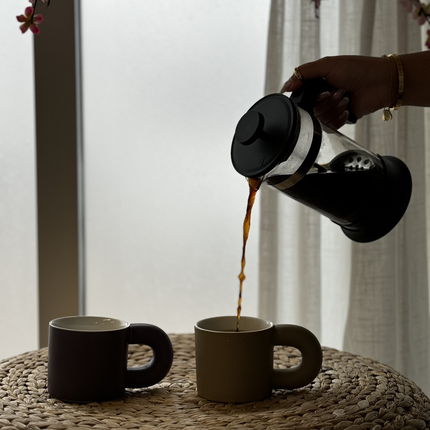 Person pouring coffee from a thermos into two black mugs on a textured surface with a blurred background.