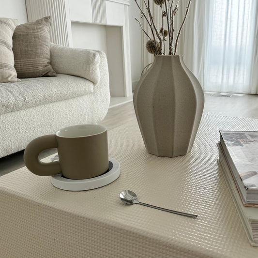 The image features a cozy setting with two ceramic mugs, one purple and one gray, placed on a table alongside a heated coaster and a silver spoon. The scene includes a decorative vase with pink cherry blossoms, adding a touch of elegance and tranquility to the atmosphere.