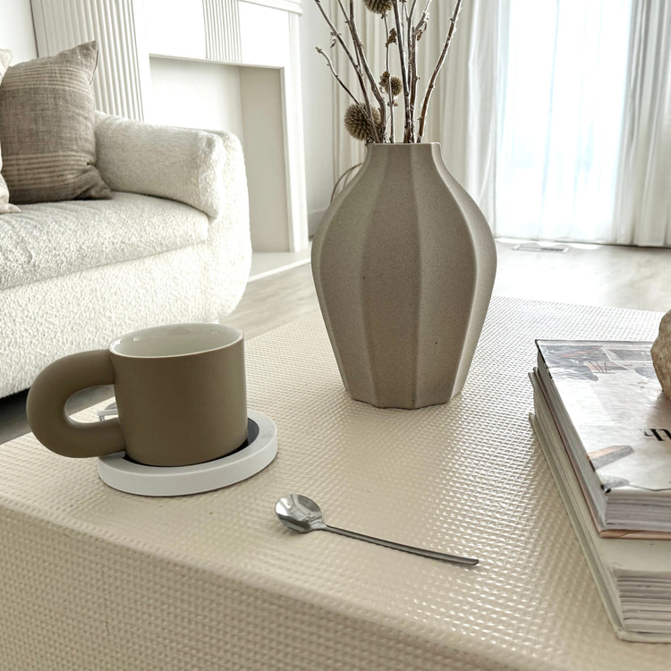 Beige mug, vase, and spoon on a textured surface with a couch and window in the background
