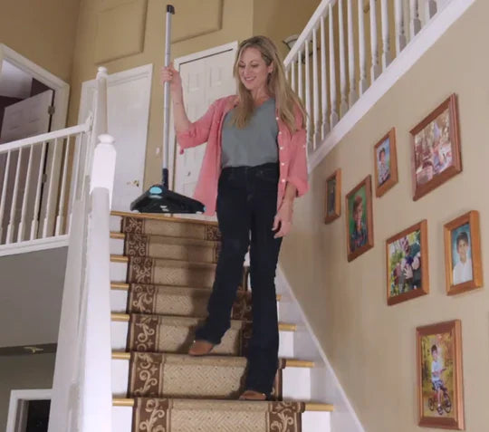Woman cleaning a staircase with a duster, surrounded by family photos on the wall.