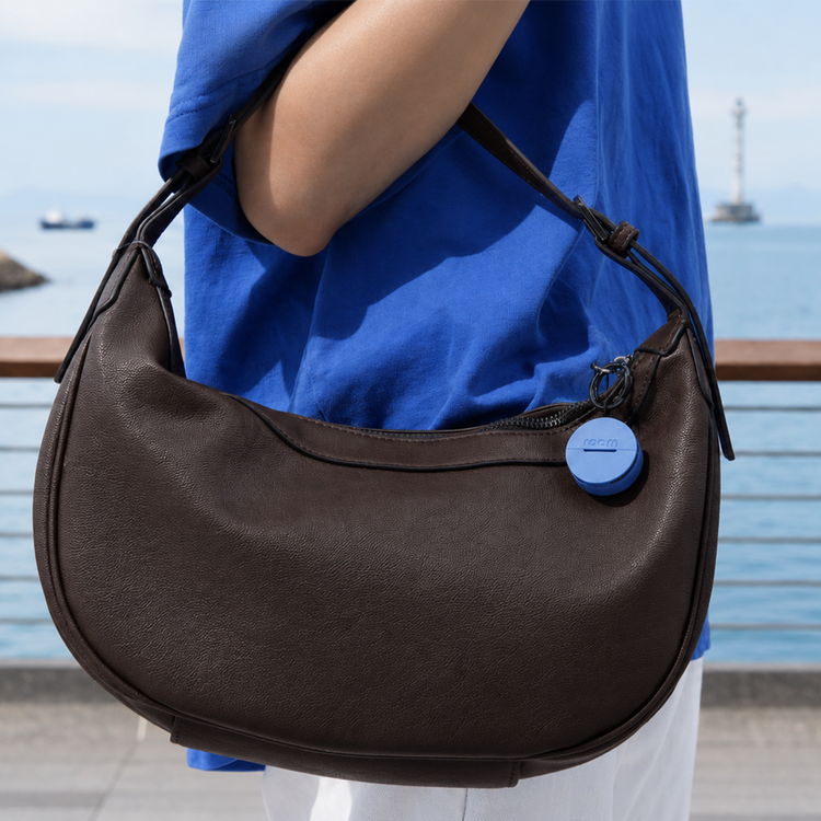 Person holding a brown handbag with a blue button, standing by a waterfront.