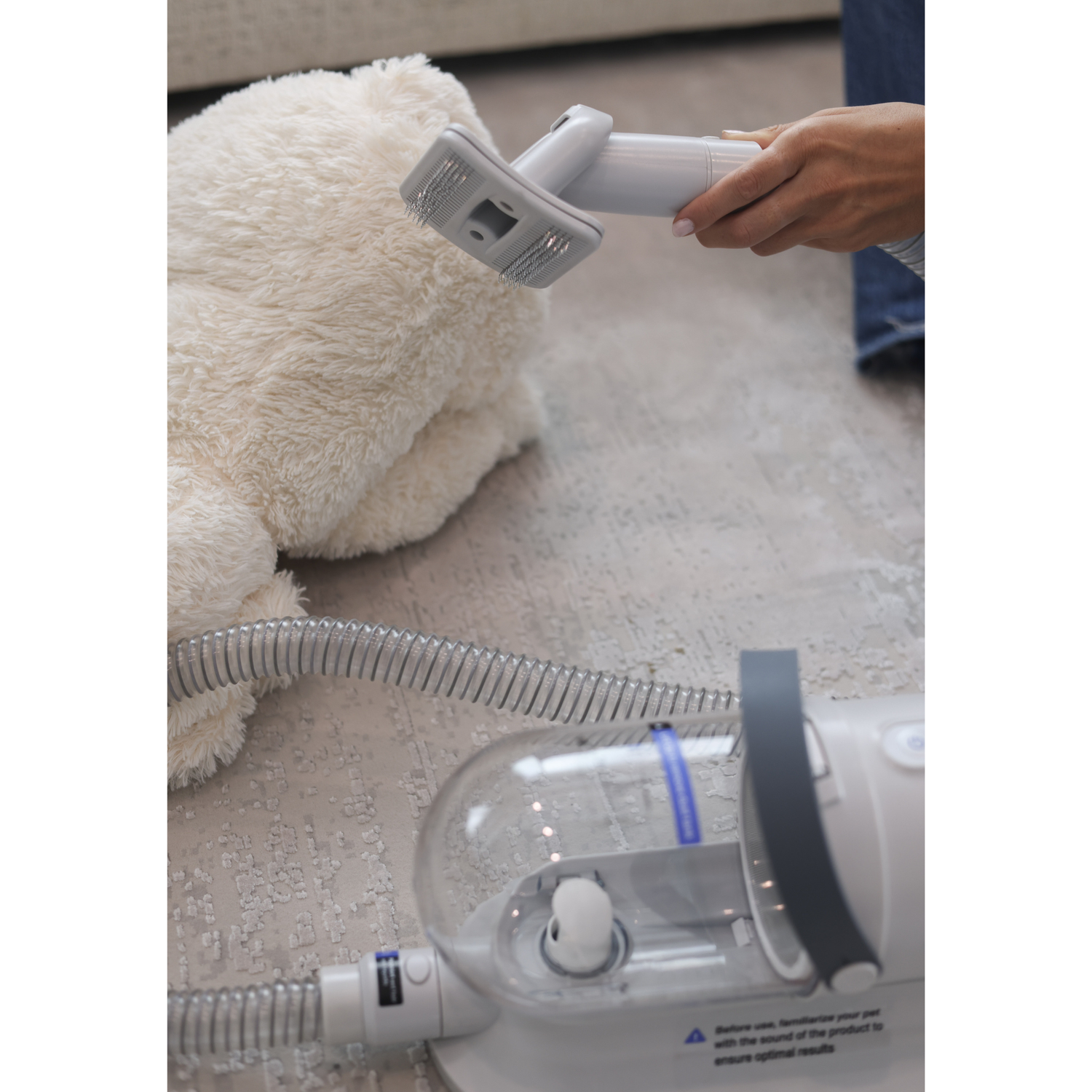 Person using a handheld device on a fluffy white dog with a vacuum cleaner on the floor.